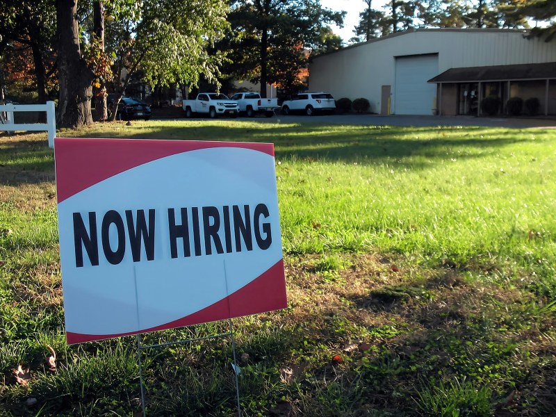 A red and white sign that reads Now Hiring in the grass outside a business.